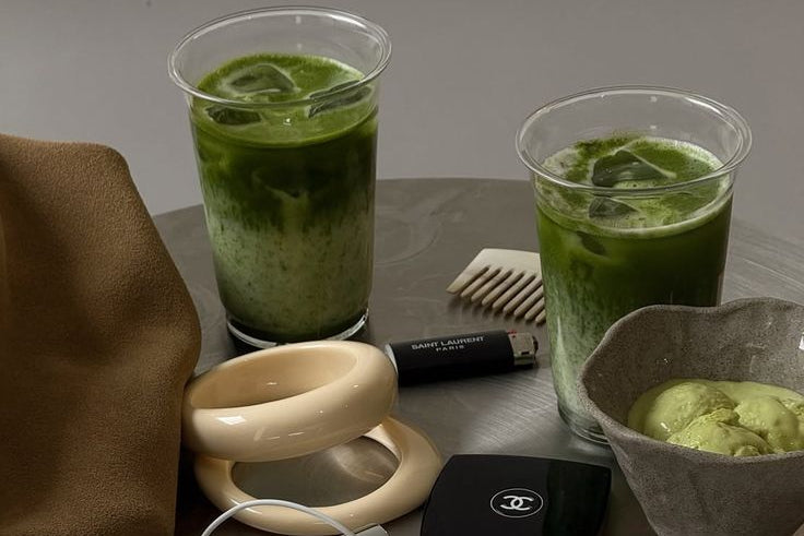Green smoothie in a clear cup with a bowl of green food, Chanel earphones, and a comb on a gray surface.